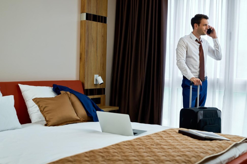a businessman communicating on a mobile phone while standing with a travel bag in a hotel room