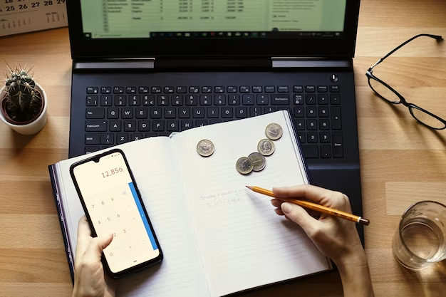 Person holding a smartphone in one hand and a pencil in the other hand, with a notebook and laptop beneath the notebook, and coins resting on top